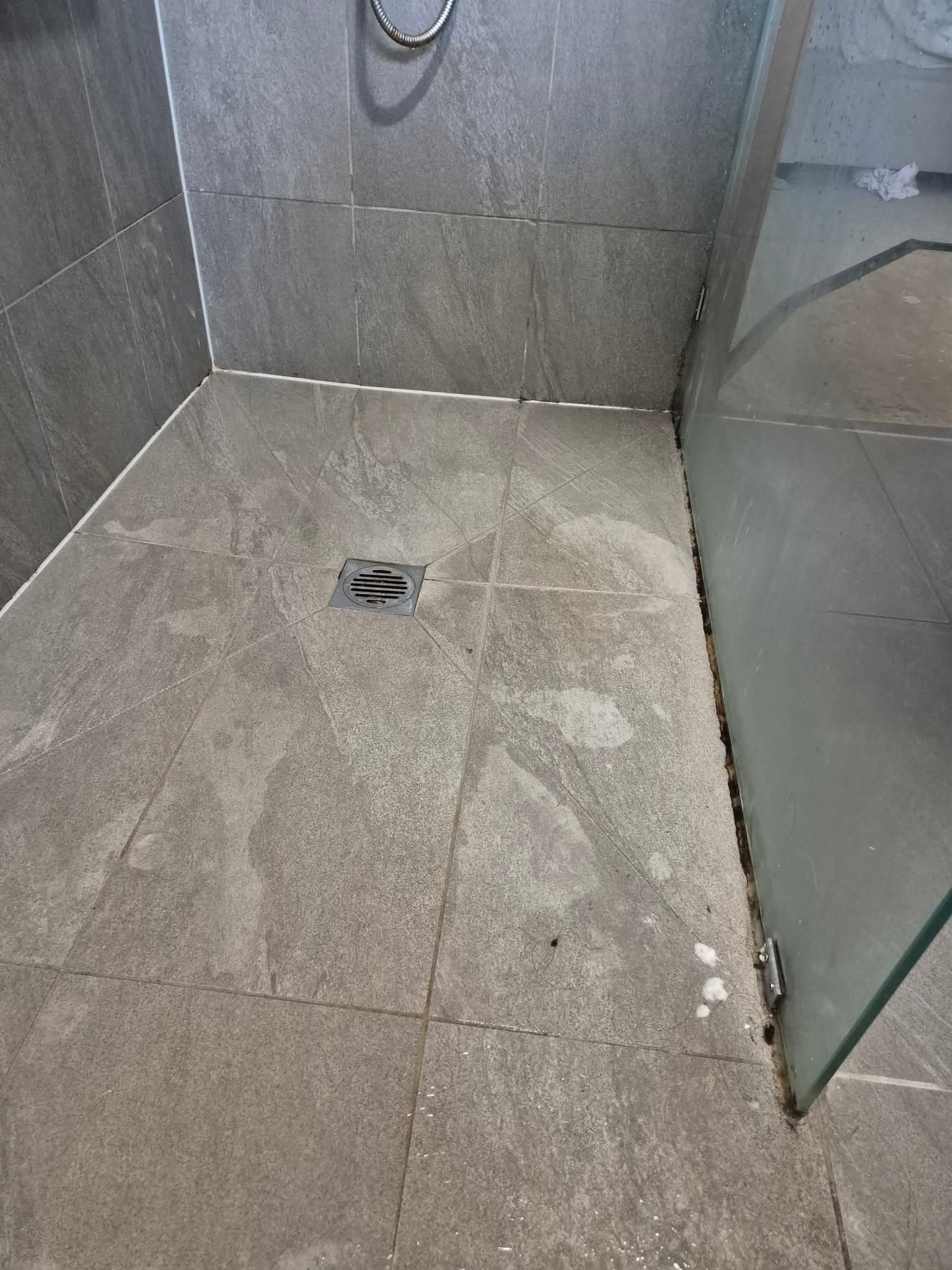 Corner of a shower with grey tiles and a glass door with visible grime along the edges.