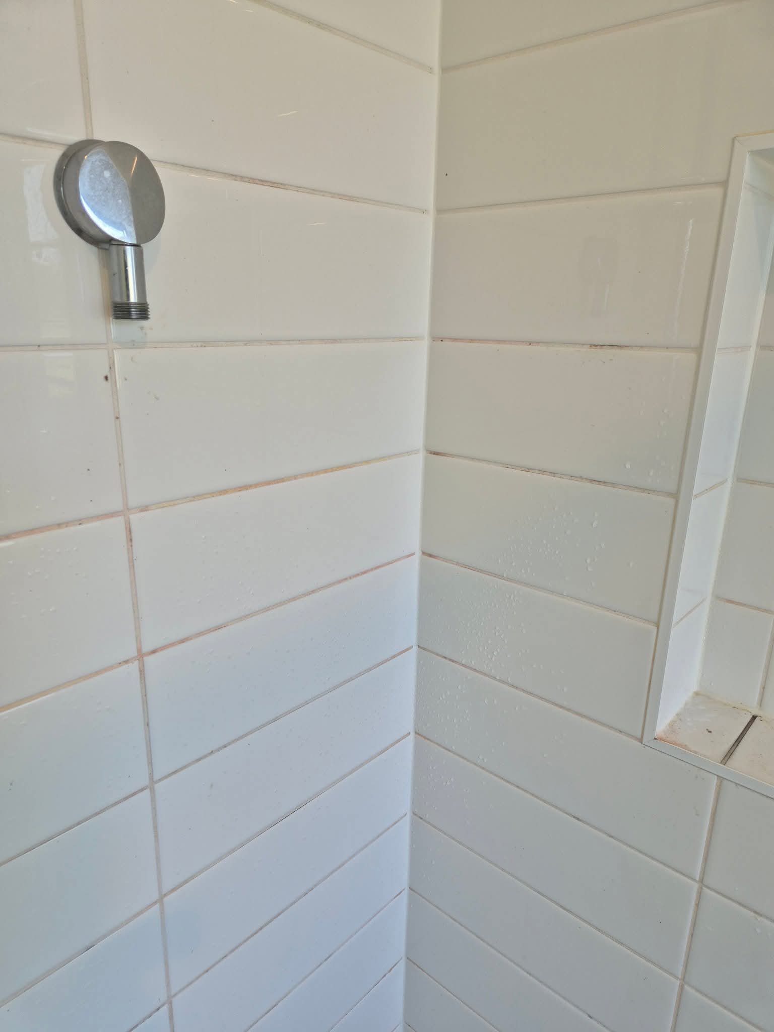 Corner of a tiled shower with a wall-mounted shower head fixture and a recessed shelf.